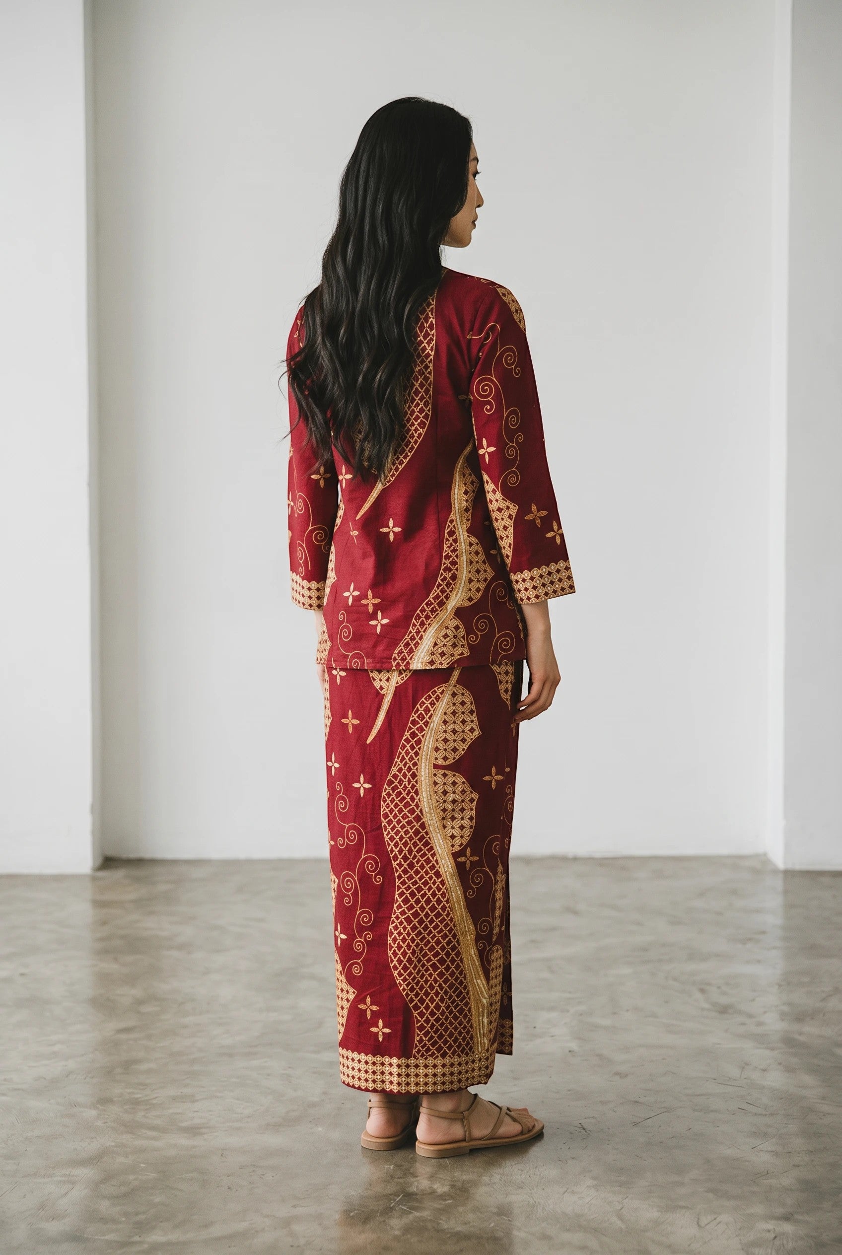 A woman wearing a maroon PARCA cotton top and matching maxi sarung with traditional gold batik-style patterns.