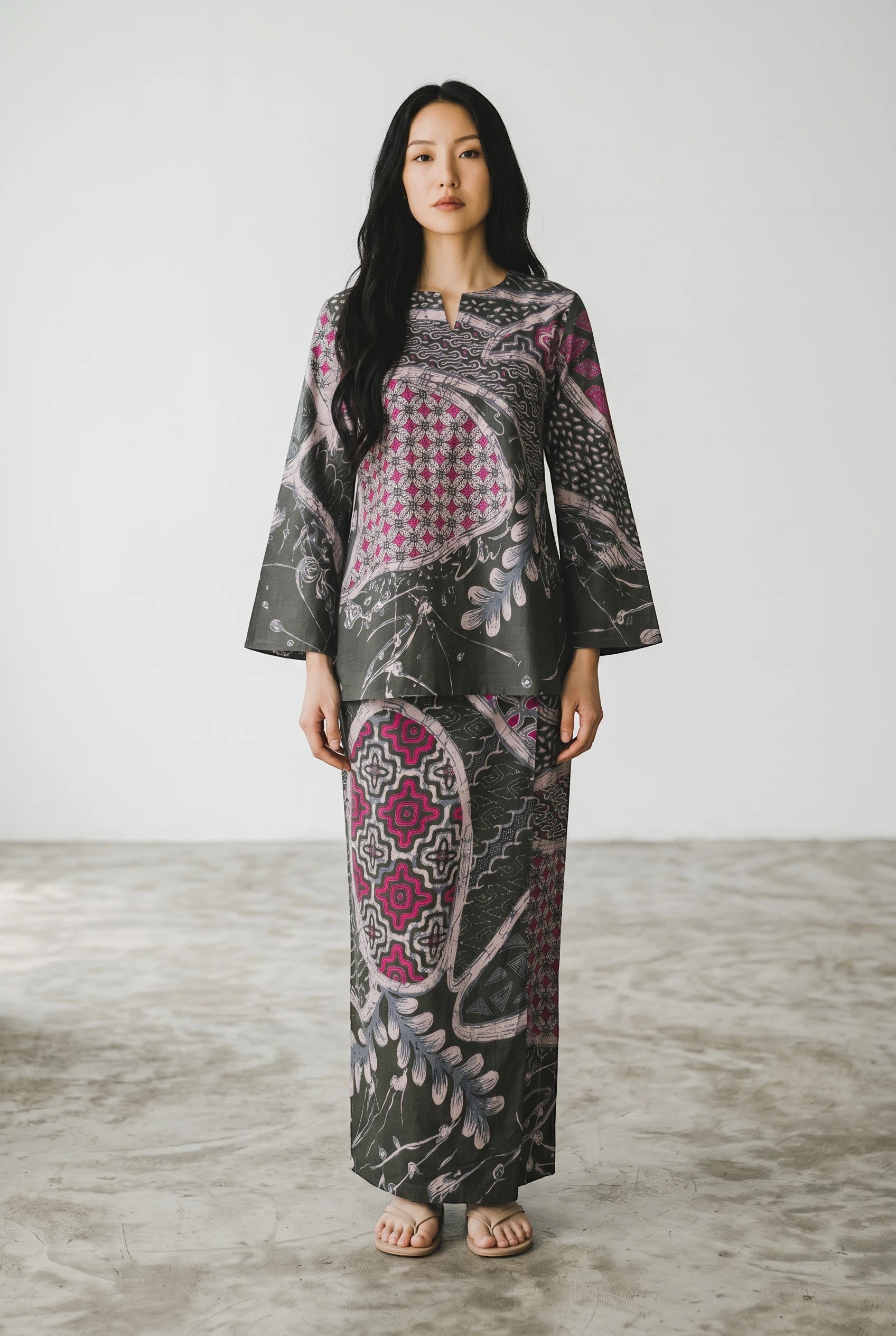 Model wearing a PARCA Munawara two-piece set in dark grey with pink batik-inspired floral and geometric patterns.